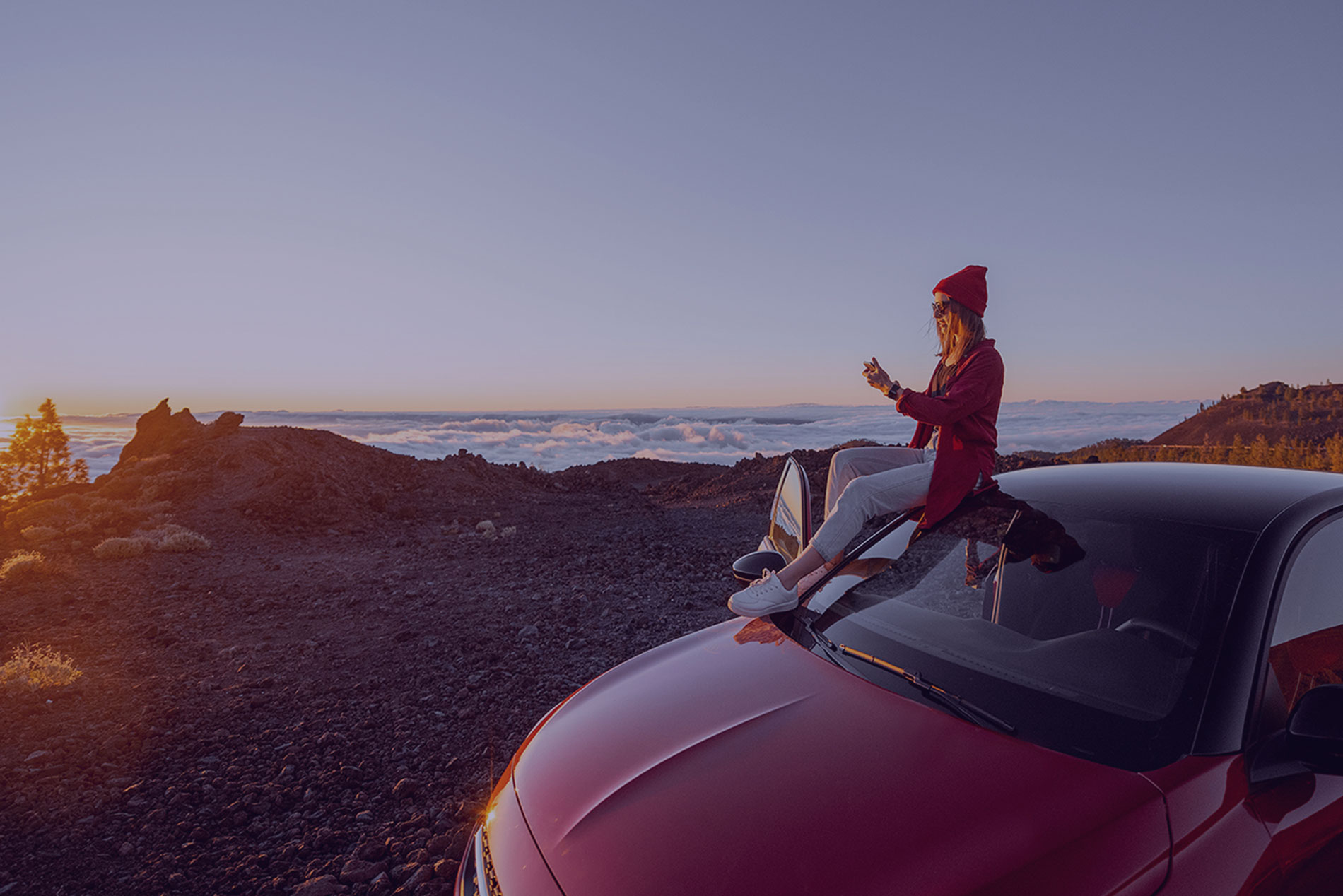Woman sitting on a red car above the clouds at sunset